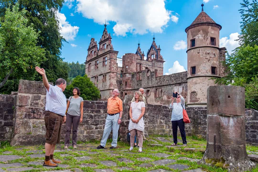 Kloster-Hirsau_Klosterführung_Foto-avmediafactory-Gert-Tetzner_Quelle-Stadt-Calw_1080 Ein Reiseleiter gestikuliert, während er mit einer Gruppe von fünf Touristen in einer alten Burg mit kunstvoller Steinarchitektur spricht. Die Szene spielt im Freien an einem hellen Tag mit blauem Himmel und Wolken im Hintergrund.