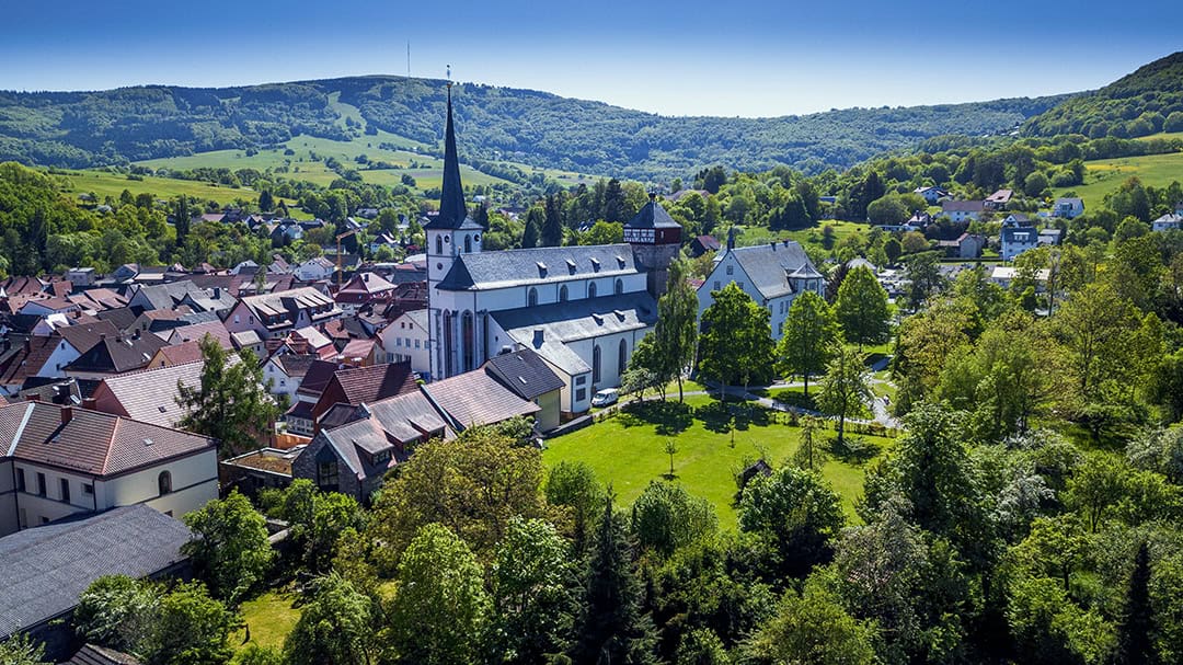 Luftaufnahme-Pfarrgarten-Stadtpfarrkirche-Stadtturm-Rentamt_1080 Luftaufnahme eines malerischen Dorfes in einem üppigen Tal. Eine markante Kirche mit hohem Turm steht inmitten dicht gedrängter Häuser mit roten Dächern. Das Dorf ist von sanften grünen Hügeln und dichten Wäldern unter einem klaren blauen Himmel umgeben.