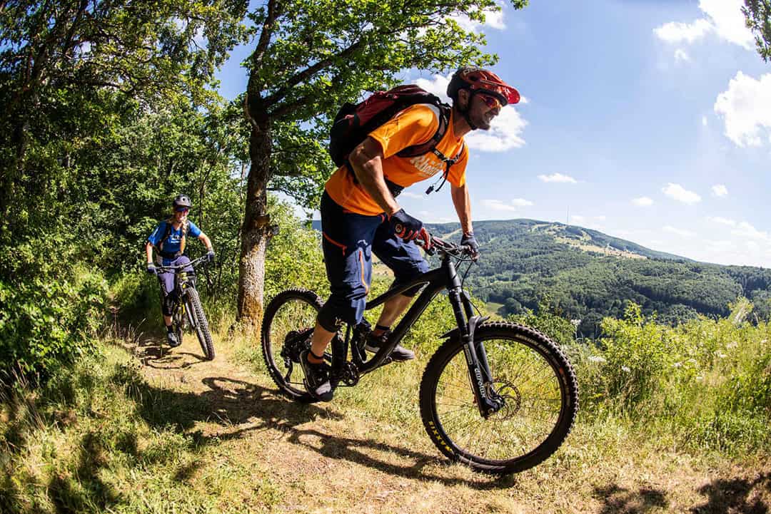 Mountainbiken-Bischofsheim-Kreuzberg_1080 Zwei Mountainbiker fahren auf einem malerischen Trail durch einen üppigen, grünen Wald. Der vordere Radfahrer trägt ein orangefarbenes Shirt und einen roten Helm, gefolgt von einem weiteren Radfahrer. Die Sonne wirft helles Licht auf die Straße, und im Hintergrund sind sanfte Hügel unter einem blauen Himmel zu sehen.