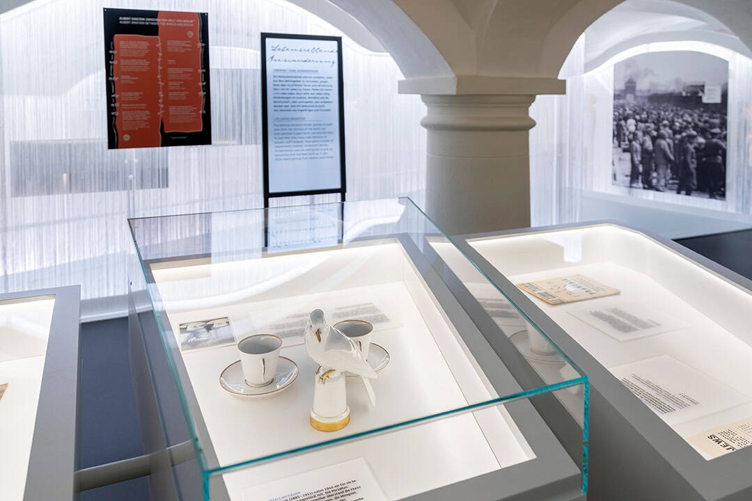 Die-Einsteins_Emigration_Nadja-Wollinsky_1080 Museumsausstellung mit historischen Dokumenten und Artefakten in Glasvitrinen. Enthält ein Tassenset mit einer weißen Taubenfigur, begleitet von beschreibenden Tafeln und einem großen historischen Foto im Hintergrund. Elegante Bogenarchitektur umrahmt die Ausstellung.