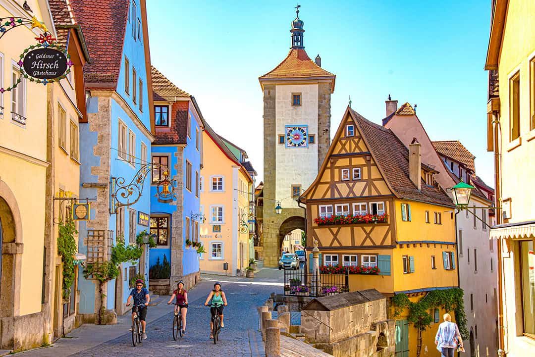 Radfahrer-Rothenburg-Thomas-Weller_1080 Eine malerische Straße in Rothenburg ob der Tauber mit farbenfrohen Fachwerkhäusern und einem historischen Torturm. Drei Personen fahren an einem sonnigen Tag mit dem Fahrrad die Kopfsteinpflasterstraße entlang. Ein Schild des Gasthauses Hirsch ist zu sehen.