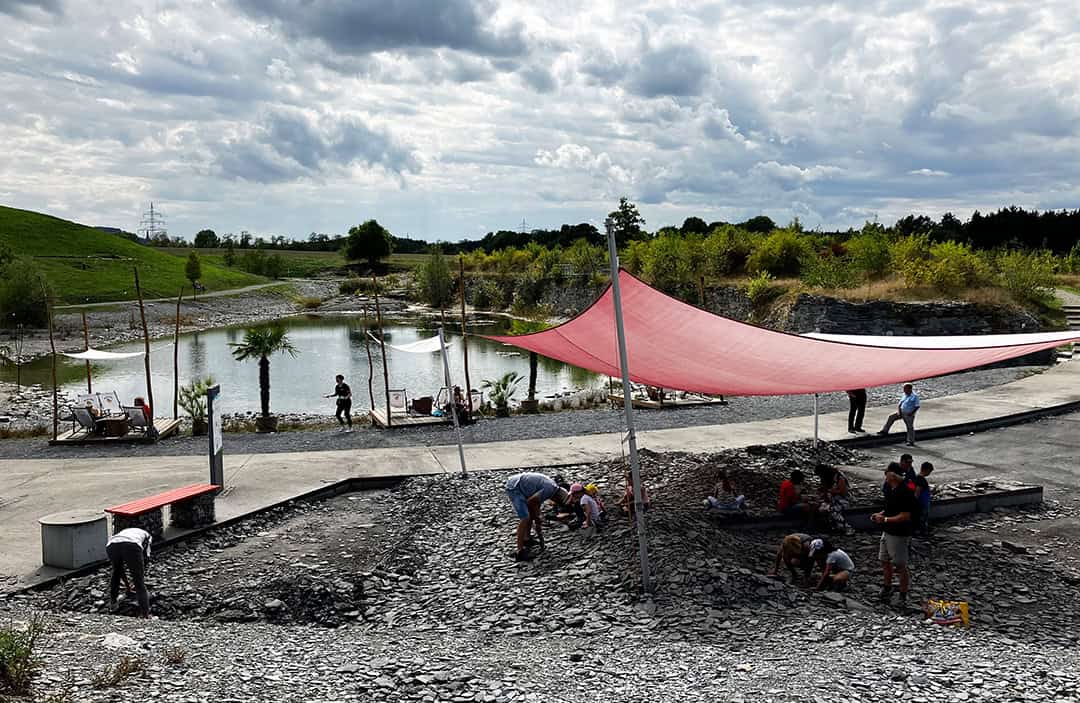 SCHIEFERERLEBNIS_Foss.Sammelplatz_1080 Menschen haben sich um ein felsiges Gebiet unter einem roten dreieckigen Baldachin versammelt. Im Hintergrund ist ein Teich mit einem kleinen Springbrunnen zu sehen, umgeben von Grün. Der Himmel ist bewölkt, und mehrere Menschen scheinen zu graben oder die Felsen zu erkunden.