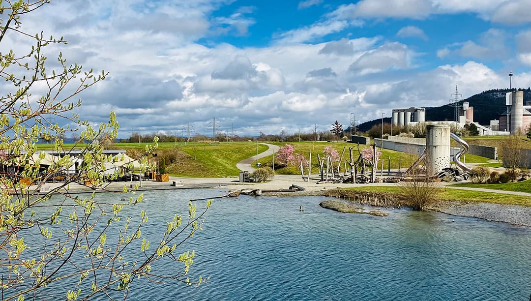 SE_BlickSchiefersee_1080 Malerische Landschaft mit einem ruhigen See im Vordergrund, einem gewundenen Pfad, der durch eine Grasfläche mit blühenden Bäumen führt, und Industrieanlagen im Hintergrund unter einem teilweise bewölkten Himmel.