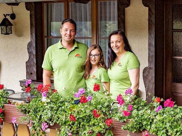 csm_Alisehof_Fam_Schreier_600 Drei Personen, zwei Erwachsene und ein Kind, stehen auf einem mit bunten Blumen geschmückten Balkon. Sie tragen passende grüne Hemden und lächeln. Der Balkon ist mit Holzelementen versehen, dahinter ist ein Fenster mit Vorhängen zu sehen.