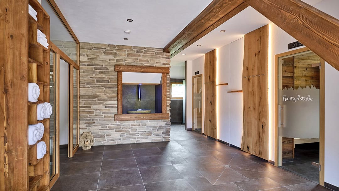 csm_Alisehof_Sauna_01_1080 Spa-Innenraum mit Stein- und Holzwänden, Schieferboden und warmer Beleuchtung. Handtücher liegen links in einem Holzregal. Rechts befindet sich eine Glastür mit dem Schriftzug „Bruggerstube“. Ein kleines Fenster gibt den Blick auf ein beleuchtetes Wasserspiel frei.