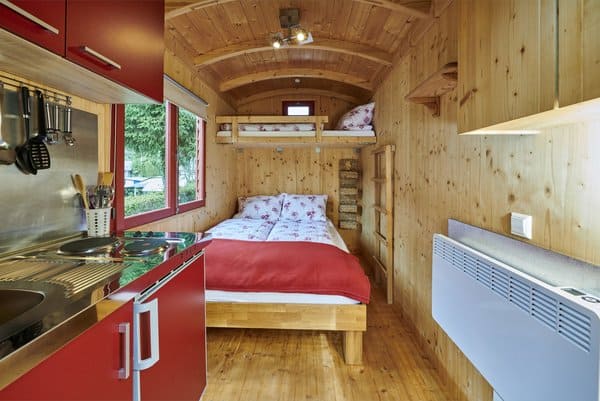 csm_Alisehof_Schaeferwagen_600 Gemütliches Interieur eines kleinen Holzhauses mit einem Doppelbett unter einem Einzelbett. Die kleine Küche links ist mit roten Schränken ausgestattet. Der Raum ist mit Holzvertäfelungen ausgestattet, und an der rechten Wand ist eine Heizung angebracht. Ein Fenster gibt den Blick ins Grüne frei.