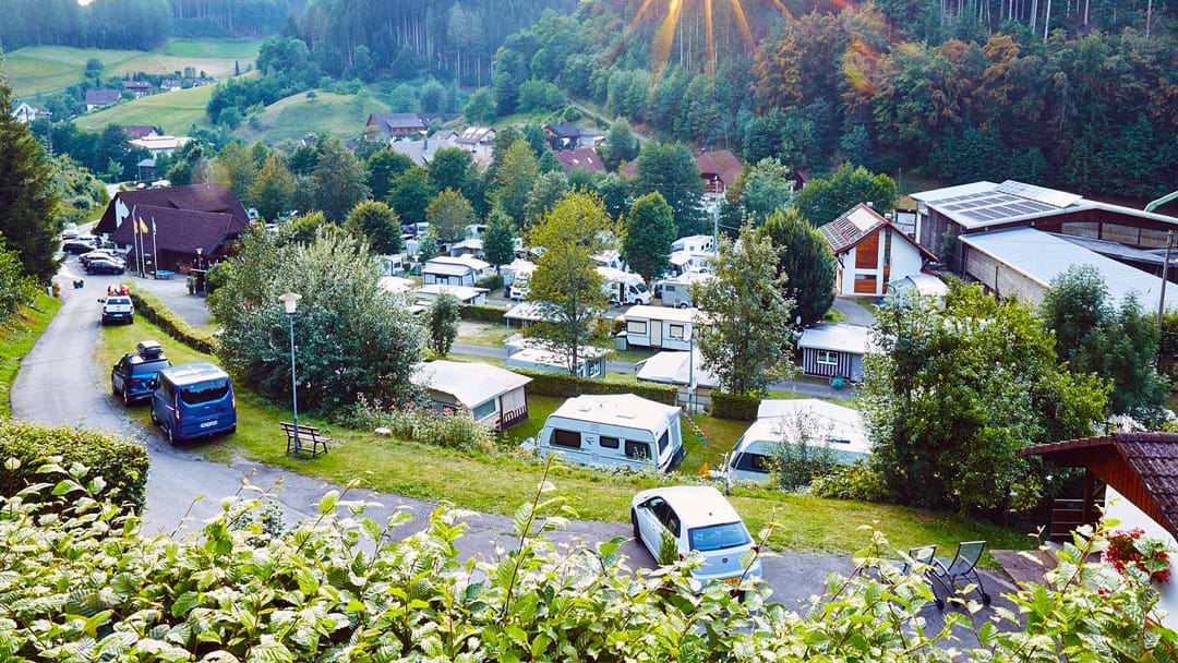 csm_Alisehof_camping_1080 Ein malerischer Campingplatz mit mehreren Wohnwagen und Zelten inmitten von Bäumen. Autos parken entlang einer kurvenreichen Straße, und ein Gebäude mit Solarmodulen ist sichtbar. Sonnenlicht fällt durch die Baumkronen und erhellt die üppige Landschaft.