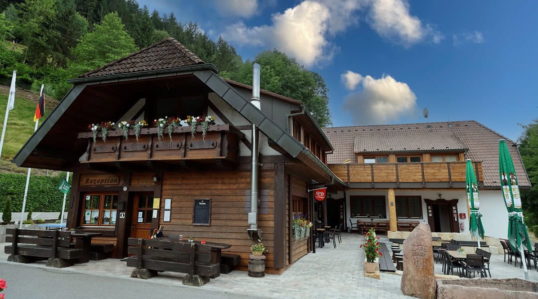 csm__Reception_1080 Eine charmante Holzhütte mit steilem Dach und blumengeschmücktem Balkon. Sie bietet Sitzgelegenheiten im Freien, ausländische Flaggen und grüne Sonnenschirme. Bäume und ein wolkenloser blauer Himmel bilden die Kulisse.