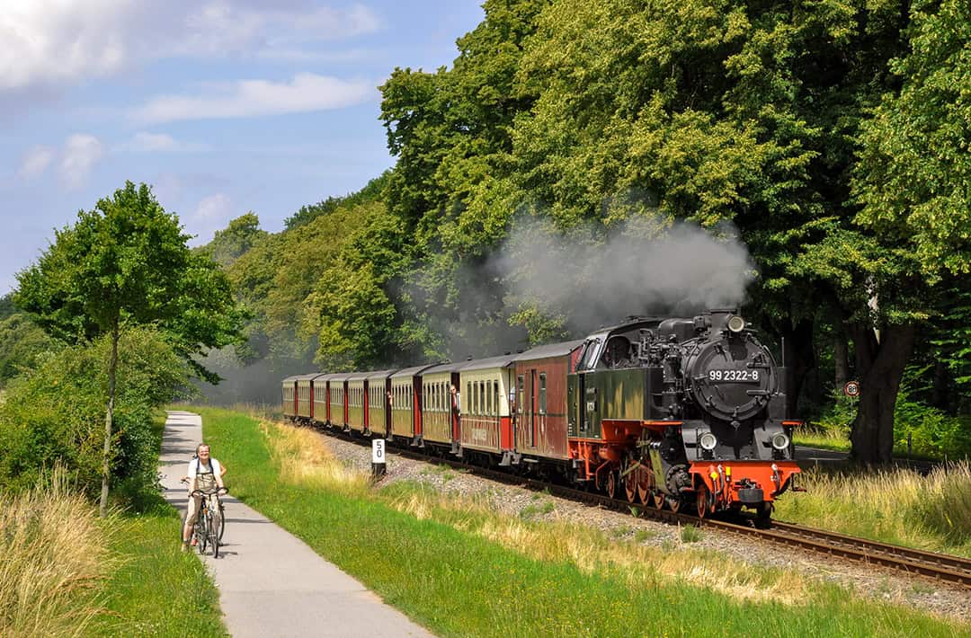 Eine Dampflokomotive fährt einen üppig grünen Weg entlang und stößt Rauch aus. Eine Person in Freizeitkleidung radelt den Weg entlang, umgeben von hohem Gras und dichten Bäumen. Der Himmel darüber ist teilweise bewölkt.