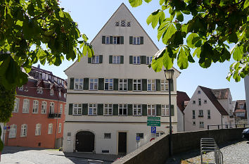 Ein weißes, mehrstöckiges historisches Gebäude mit dreieckigem Dach und grünen Fensterläden steht unter sonnigem Himmel. Bäume mit grünen Blättern rahmen den oberen Bildrand ein. Die umliegenden Gebäude haben farbenfrohe Fassaden. Eine Straßenlaterne und Verkehrsschilder sind sichtbar.