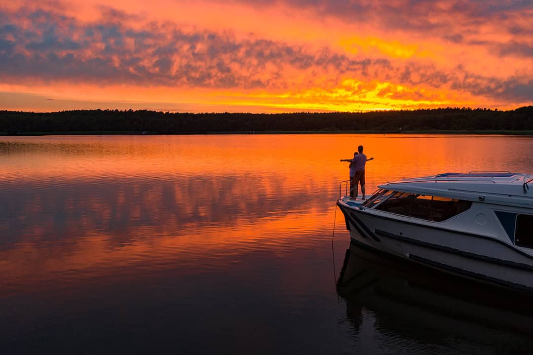 le-Boat_Holger-Leue_Romantik_1080 Eine Person steht auf einem angedockten Boot und betrachtet einen leuchtenden Sonnenuntergang über einem ruhigen See. Der Himmel wechselt von tiefem Orange zu Gelb und spiegelt sich wunderschön auf der Wasseroberfläche.