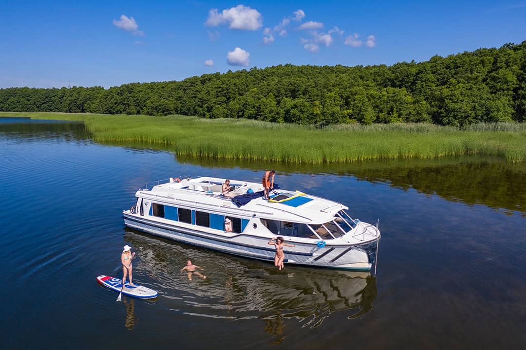 le-boat_Holger-Leue_Fürstenberg_1080 Ein weißes Boot liegt auf einem ruhigen See, umgeben von üppigen grünen Bäumen, vor Anker. Menschen entspannen sich auf dem Boot und schwimmen in der Nähe. Eine Person steht auf einem Paddleboard und genießt den sonnigen Tag und den klaren blauen Himmel.