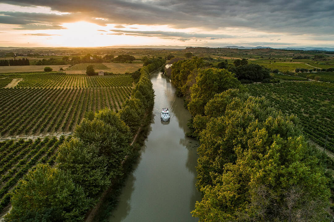 le-boat_Holger-Leue_Kanaele_1080 Luftaufnahme eines Bootes, das einen schmalen Fluss befährt, der von üppigen Weinbergen und Bäumen gesäumt ist. Die Landschaft erstreckt sich unter einem bewölkten Himmel bis in die fernen Hügel. Die Sonne geht am Horizont unter und taucht die Szene in einen warmen Schein.
