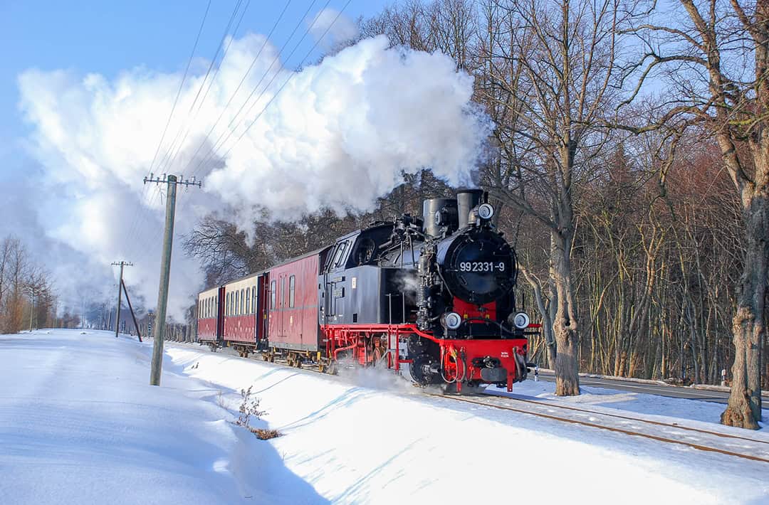 Eine schwarze Dampflokomotive mit roten Akzenten fährt auf schneebedeckten Gleisen und zieht kastanienbraune Personenwagen. Dicker Dampf quillt aus ihrem Schornstein. Kahle Bäume säumen den Hintergrund unter einem klaren blauen Himmel.