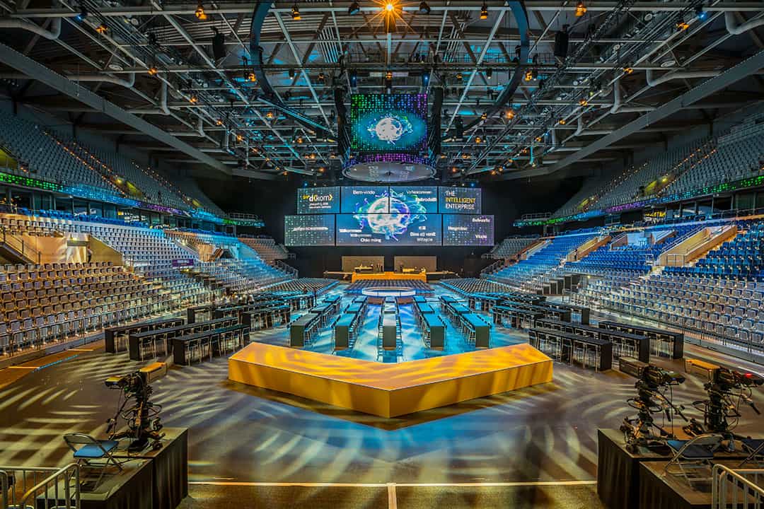 Geräumige Indoor-Arena für ein E-Sport-Event mit zahlreichen Monitoren und Gaming-Stationen, die in einem zentralen Sechseck angeordnet und von Kameras umgeben sind. Große Bildschirme zeigen Eventgrafiken, und rund um den Veranstaltungsort sind Sitzreihen zu sehen.