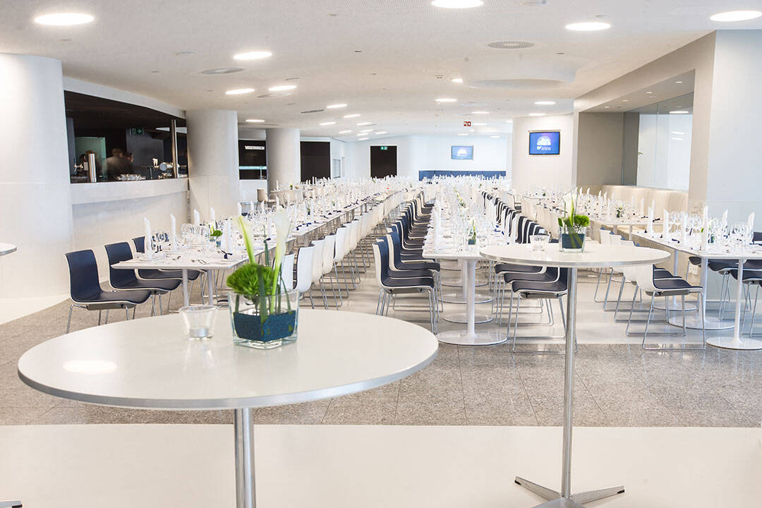 Moderner Speisesaal mit langen, gedeckten Tischen für eine Veranstaltung. Die Tischreihen sind mit weißen Tischdecken, Glasgeschirr und grünen Pflanzen dekoriert. Ein kleiner runder Tisch im Vordergrund steht mit einer Pflanze und einem Glas. Helles und großzügiges Ambiente.