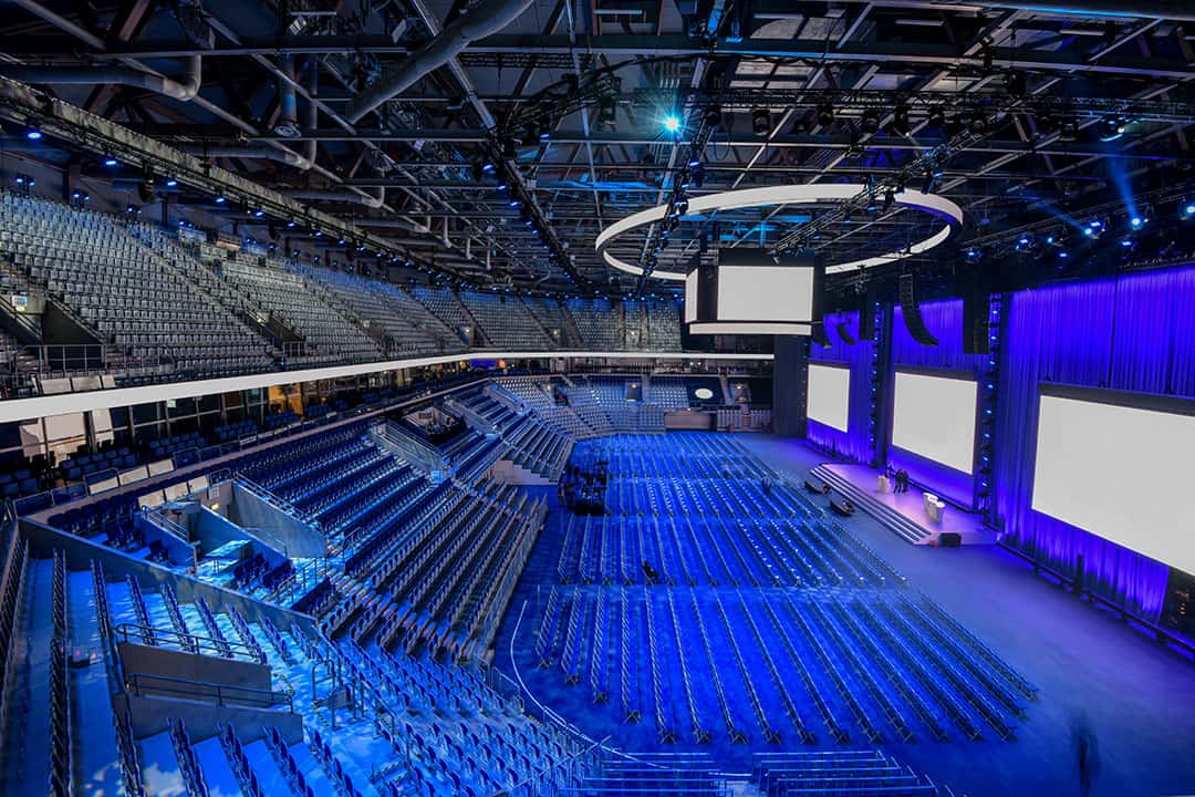 Eine große, moderne Indoor-Arena mit Sitzreihen und einer Bühne mit mehreren Großbildschirmen. Die blaue Beleuchtung des Veranstaltungsortes sorgt für eine lebendige Atmosphäre. Der Raum ist leer, was die enorme Sitzplatzkapazität unterstreicht.