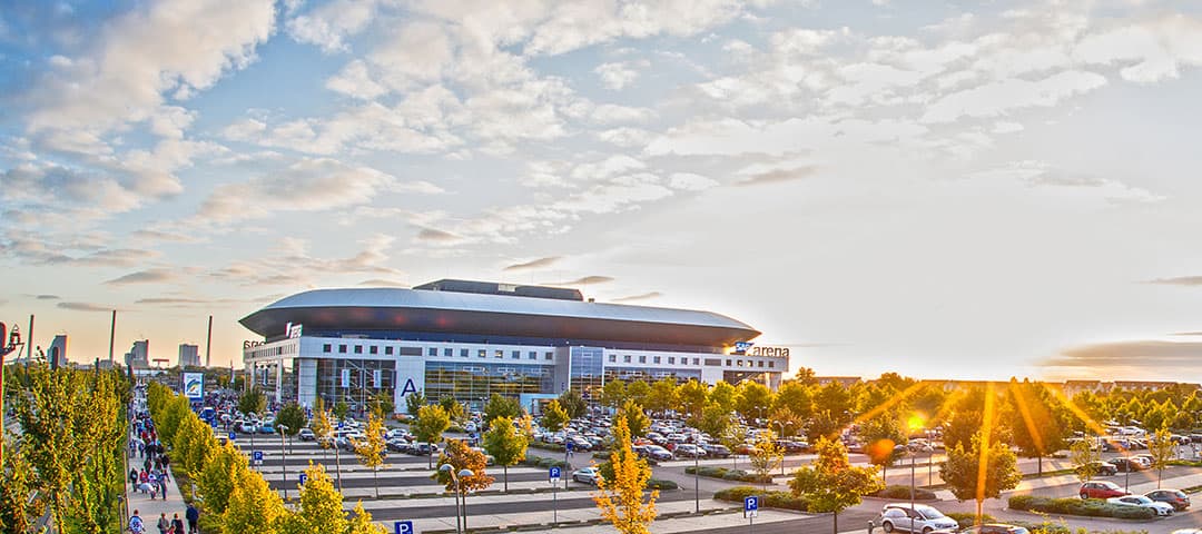 SAP-Arena-Panorama_Tobias-Schwerdt_1080 Eine große, moderne Arena mit geschwungenem Dach ist von einem Parkplatz umgeben. Menschen gehen auf baumgesäumten Wegen zum Eingang. Die Sonne geht am Horizont unter und taucht die Szene in einen warmen Schein. Am Himmel sind vereinzelte Wolken zu sehen.