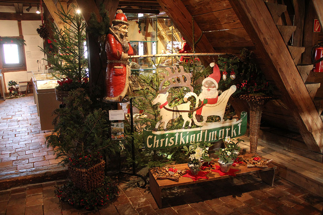 Eine festliche Innendekoration zeigt eine Weihnachtsmannfigur neben einem Schild mit der Aufschrift „Christkindlmarkt“ und einer Abbildung des Weihnachtsmanns in einem von einem Rentier gezogenen Schlitten. Tannenzweige und Dekorationen umgeben die Szene auf einem rustikalen Ziegelboden.
