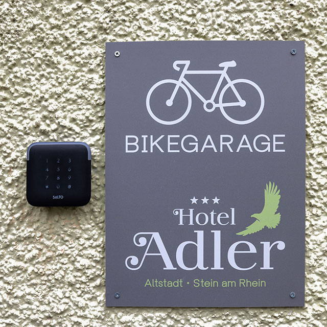 Eine strukturierte Wand mit einem Schild mit einem Fahrradsymbol und den Worten BIKEGARAGE und Hotel Adler, Altstadt, Stein am Rhein neben einem Zugangsgerät mit Tastatur.
