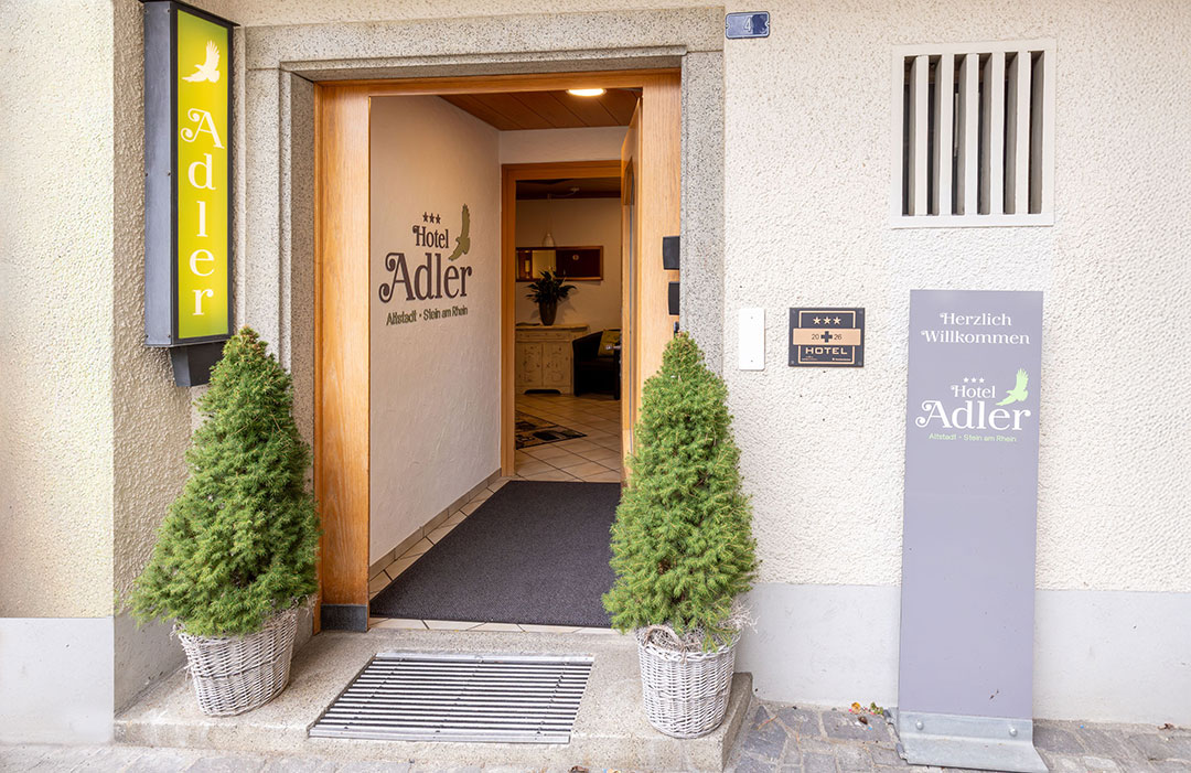 Eingang des Hotels Adler mit offener Holztür, zwei Topfsträuchern und Schildern mit Hotelinformationen. Das Wandschild zeigt die Drei-Sterne-Bewertung. Das Willkommensschild lautet „Herzlich Willkommen“. Beigefarbene Fassade mit einem kleinen Fenster darüber.