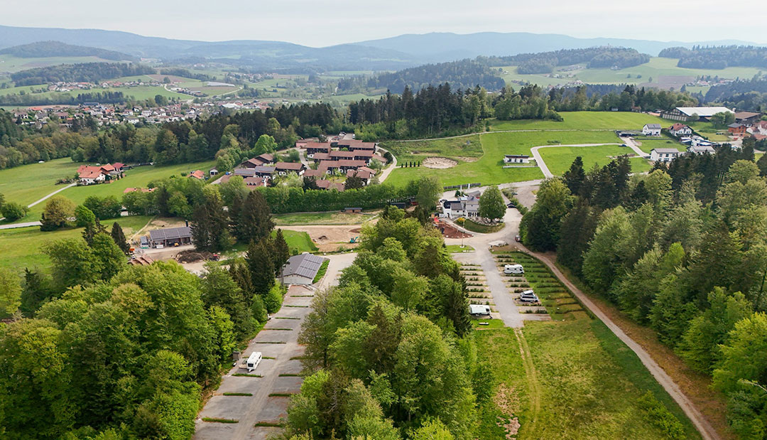 BWC_Luftbild_1080 Luftaufnahme einer ländlichen Landschaft mit einer Kleinstadt, umgeben von grünen Feldern und Wäldern. Im Vordergrund sind parkende Autos und eine Straße zu sehen. Im Hintergrund sind sanfte Hügel und ferne Berge zu sehen.