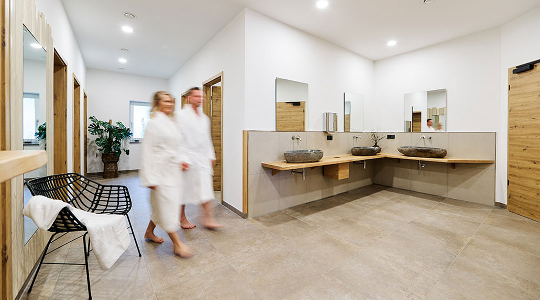 Campingresort_Bayerwald_Sanitär_1080 Zwei Personen in weißen Bademänteln gehen durch ein geräumiges, modernes Badezimmer mit beigefarbenen Fliesen und Holzakzenten. Der Raum verfügt über zwei Steinwaschbecken auf einer Holzplatte, Spiegel und einen schwarzen Drahtstuhl mit Handtuch. Eine Topfpflanze schmückt die Ecke.