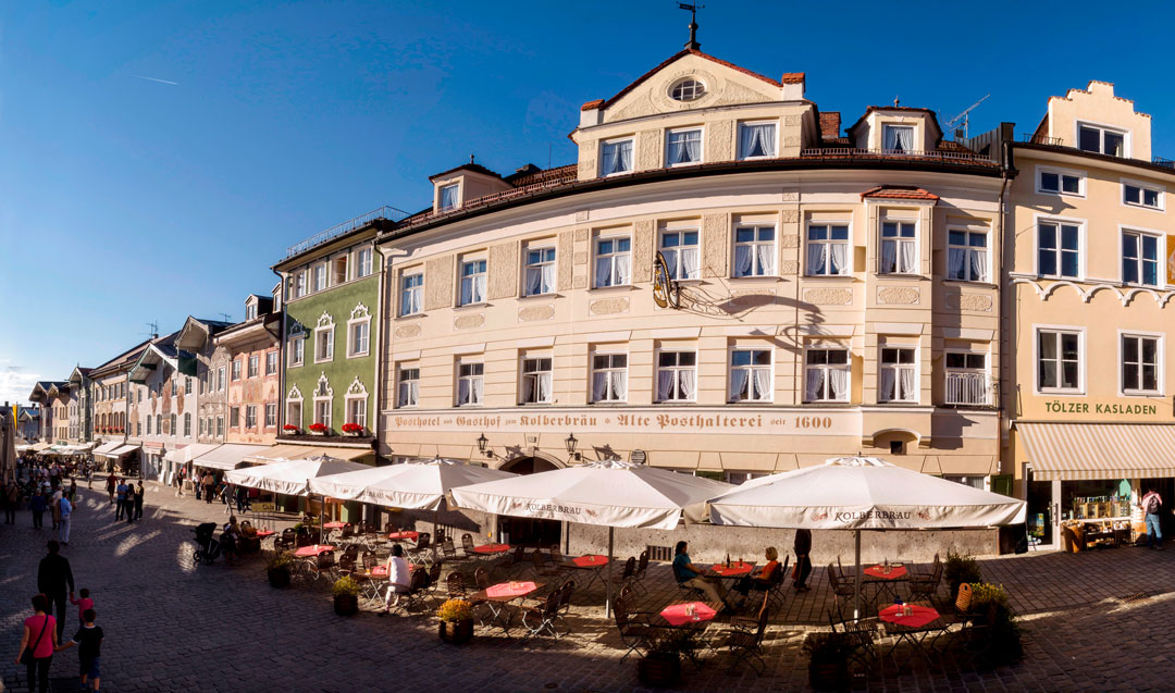 Eine lebendige Straßenansicht in Bad Tölz, Deutschland, mit farbenfrohen historischen Gebäuden unter einem strahlend blauen Himmel. Menschen schlendern und speisen an Tischen im Freien mit weißen Sonnenschirmen entlang der Kopfsteinpflasterstraße. Die Szene ist geschäftig und doch charmant.
