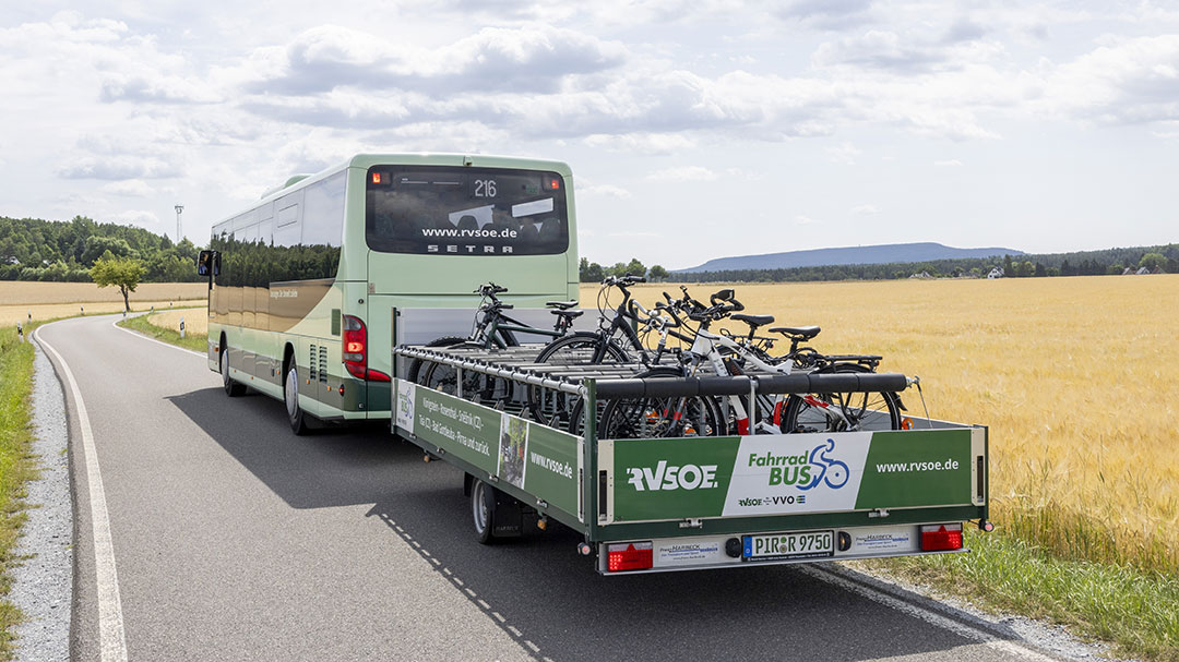 Ein grüner Bus mit einem Anhänger voller Fahrräder fährt eine Landstraße entlang. Goldene Felder und Hügel zeichnen sich am sonnigen Horizont ab. Der Anhänger trägt Logos und Website-Informationen eines Fahrradbusunternehmens.