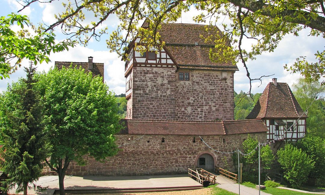 Eine mittelalterliche Steinburg mit Fachwerk im oberen Teil, umgeben von üppigen grünen Bäumen. Ein Weg führt zum Eingang, und darüber ist der blaue Himmel mit Wolken sichtbar.