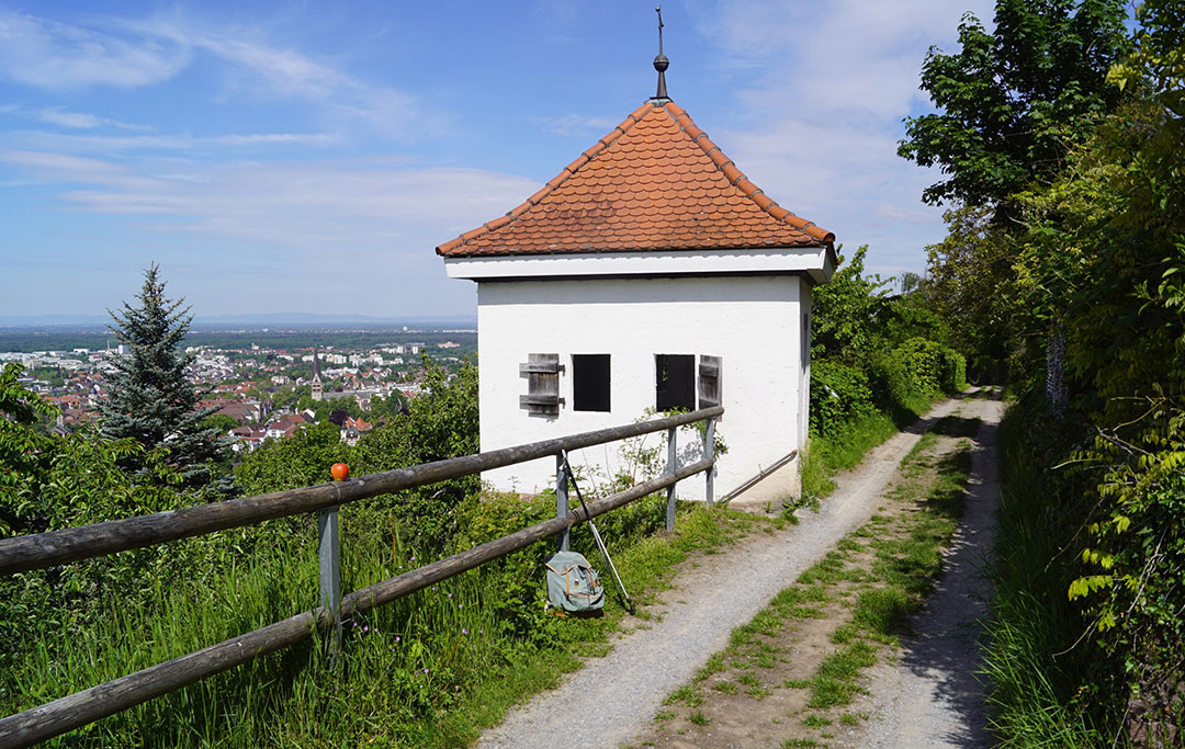 Ein kleines weißes Gebäude mit rotem Ziegeldach steht auf einem grasbewachsenen Hügel. Ein Feldweg verläuft entlang eines Holzzauns. Bäume und Grün umgeben das Gelände. Im Hintergrund blickt man auf eine Stadt und den Horizont unter blauem Himmel.