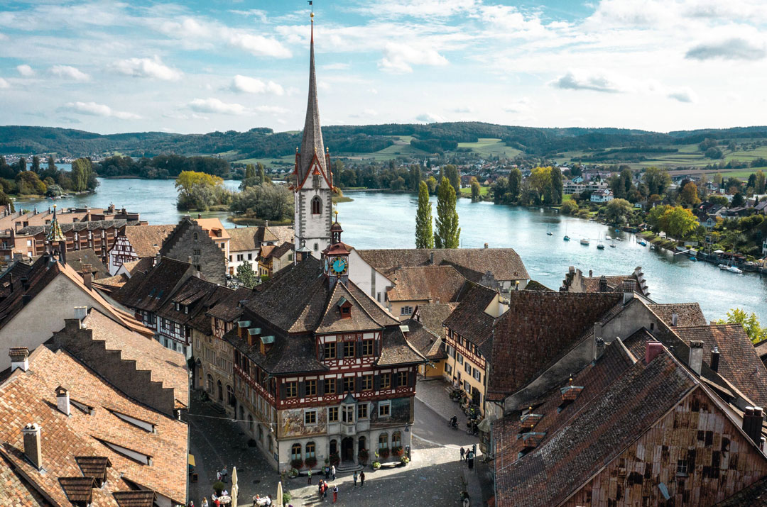 Ein malerisches Dorf, durch das sich ein Fluss schlängelt, mit einer markanten Kirche mit hohem Turm, umgeben von charmanten Gebäuden im mittelalterlichen Stil. Saftig grüne Hügel und vereinzelte Wolken prägen den Hintergrund.