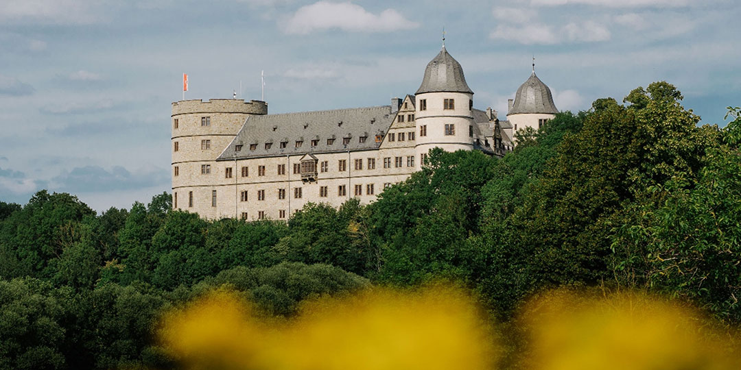 wewelsburg_1080 Eine große Steinburg mit mehreren Türmen und einer Flagge auf dem Gipfel ist teilweise von üppigen grünen Bäumen verdeckt. Im Vordergrund sind verschwommene gelbe Blumen zu sehen. Der Himmel ist blau mit vereinzelten Wolken.