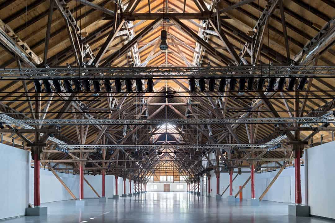 Eine geräumige, leere Industriehalle mit hoher Holzdecke und freiliegenden Balken. Der Innenraum ist mit weißen Wänden verkleidet, und von der Decke hängen Metallleuchten. Der Boden ist glatt und spiegelnd.