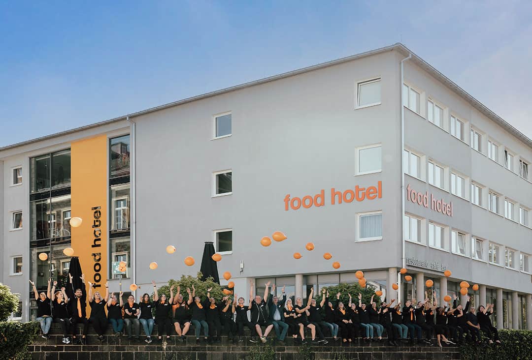 Hotel+Team_Zimpfer_1080 Eine Gruppe schwarz gekleideter Menschen sitzt vor einem grauen Gebäude mit einem Food-Hotel-Schild. Sie werfen fröhlich orangefarbene Luftballons in die Luft. Das Gebäude ist modern mit rechteckigen Fenstern und einem orangefarbenen vertikalen Banner.