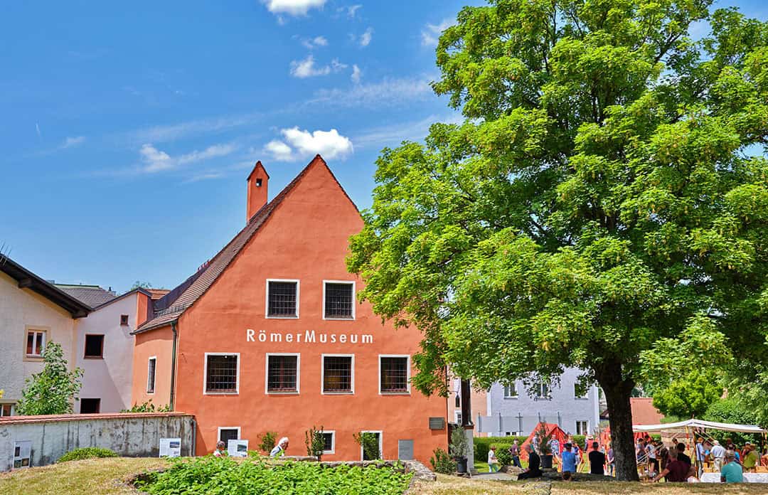 Ein sonniger Tag in einem Römermuseum. Das Gebäude ist orangefarben, hat mehrere Fenster und trägt den Schriftzug „RömerMuseum“. In der Nähe steht ein großer grüner Baum, und eine Gruppe von Menschen hat sich auf der Wiese versammelt. Der Himmel ist blau mit ein paar Wolken.