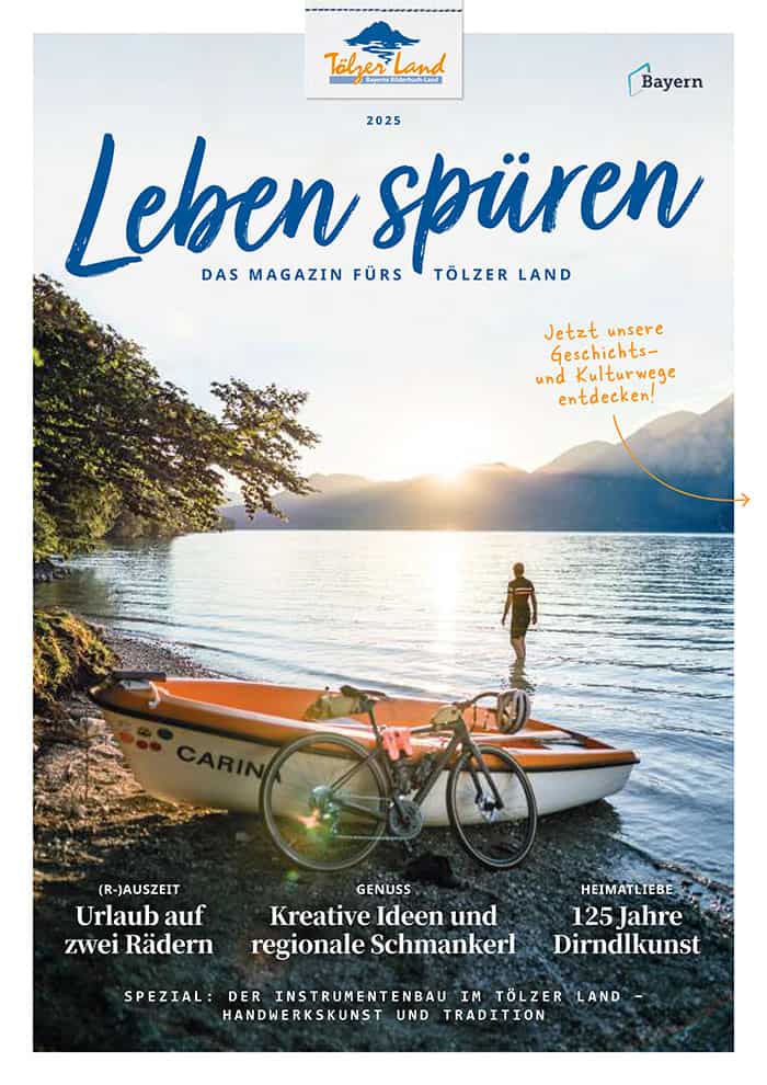 Ein Mann mit einem Holzpaddel sitzt in einem Kanu mit der Aufschrift „CARINO“ an einem See bei Sonnenuntergang. Im Hintergrund sind Berge und Bäume zu sehen. Der Text auf dem Bild enthält Zeitschriftentitel und Themen rund um Reisen und Kultur im Tölzer Land.