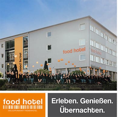 Titelseite-magazin Eine Gruppe schwarz gekleideter Menschen mit orangefarbenen Luftballons steht vor einem grauen Gebäude mit der Aufschrift „Food Hotel“. Das Gebäude hat große Fenster und ein orangefarbenes Banner. Der Text darunter lautet: „Erleben. Genießen. Übernachten.“.