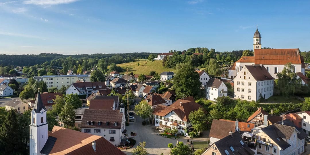 badgroenenbach_1080 Luftaufnahme eines malerischen europäischen Dorfes mit rot gedeckten Häusern, einer markanten Kirche mit hohem Kirchturm und üppigem Grün. Im Hintergrund sind sanfte Hügel unter einem klaren blauen Himmel zu sehen.