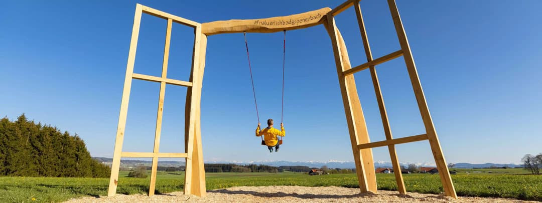 header_fruehling_1080 Eine Person in einer gelben Jacke schaukelt auf einem großen Holzrahmen auf einer Wiese, mit einem klaren blauen Himmel und fernen Bergen im Hintergrund. Der Rahmen ähnelt einem riesigen Bilderrahmen.