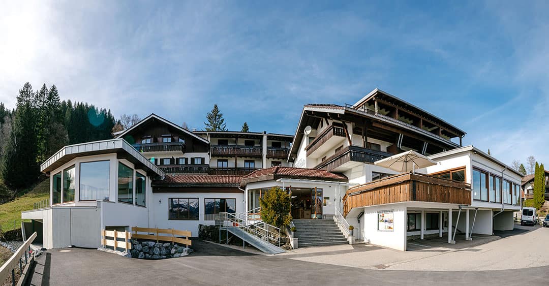 Panoramablick auf ein großes, mehrstöckiges Hotel mit Holzbalkonen und weißen Wänden unter blauem Himmel. Das Gebäude besticht durch eine Mischung aus moderner und traditioneller Architektur, umgeben von Bäumen und vor der Kulisse von Hügeln.