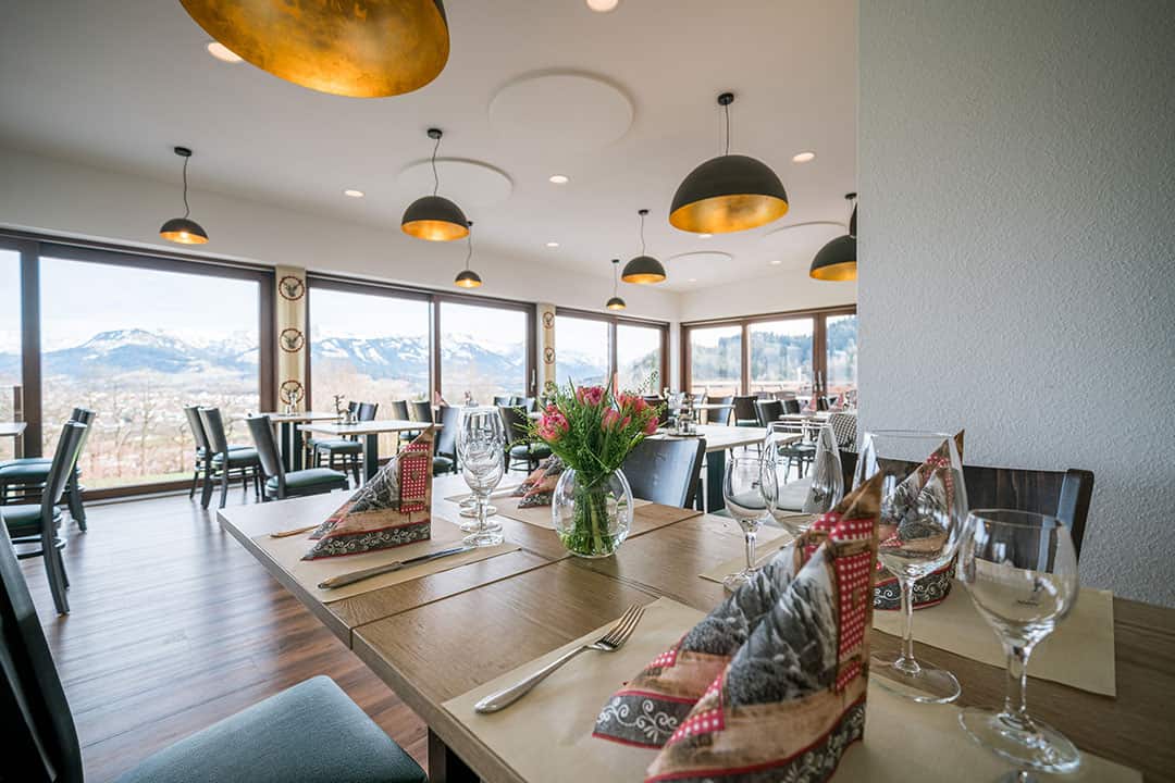 Ein elegantes Restaurant mit großen Fenstern und Blick auf die Berge. Die Tische sind mit gefalteten Servietten, Weingläsern und einem Blumengesteck gedeckt. Moderne Pendelleuchten hängen von der Decke, und der Hartholzboden verleiht dem Restaurant eine warme Atmosphäre.