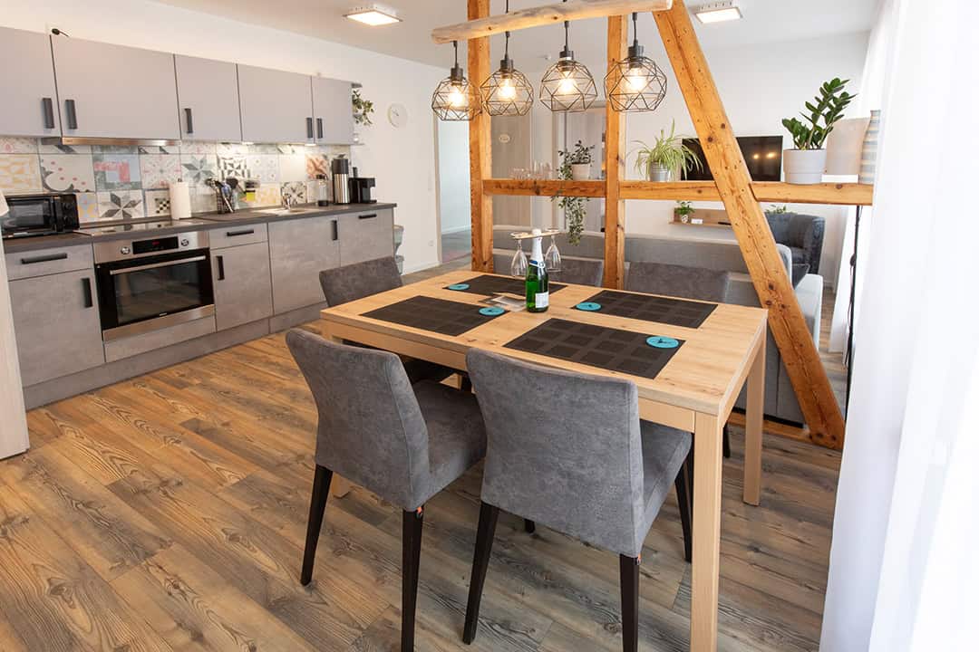 Moderne offene Küche und Essbereich mit grauen Schränken, einem hölzernen Esstisch für vier Personen, geometrischen Pendelleuchten und Holzakzenten; Pflanzen und natürliches Licht verstärken die gemütliche Atmosphäre.