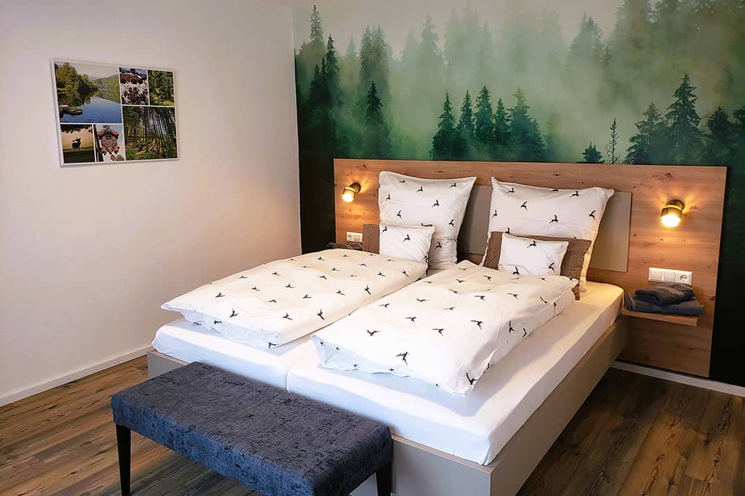 Ein gemütliches Schlafzimmer mit Doppelbett, Bettwäsche mit Vogelmuster, Holzkopfteil und zwei Wandlampen. Die Wand hinter dem Bett ist mit einem Waldgemälde geschmückt, und an der weißen Wand hängt eine Fotocollage. Am Fußende des Bettes steht eine graue Bank.