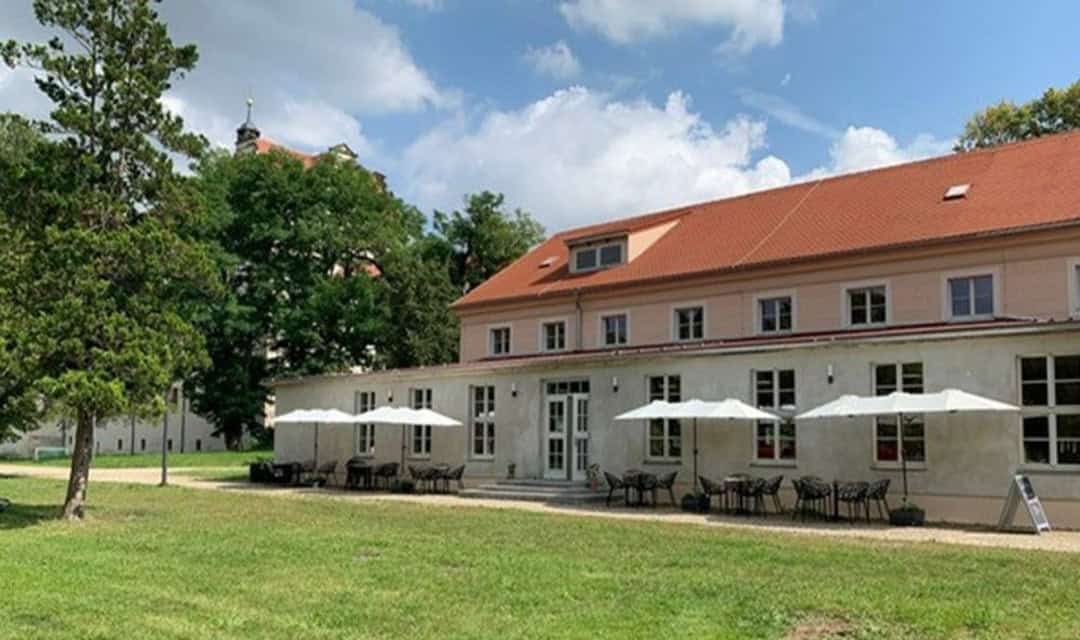 Eberhardine_Terrasse_1080 Ein helles Gebäude mit rotem Dach und Sitzgelegenheiten im Freien unter weißen Sonnenschirmen, auf einer Rasenfläche mit Bäumen und einem teilweise bewölkten Himmel im Hintergrund.