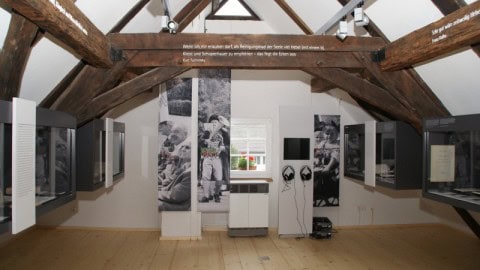 HH Dachstuhl-480x270 Ein Museumsausstellungsraum mit Holzbalken, Vitrinen an den Wänden, großen Schwarzweißfotos und einem kleinen Fenster am anderen Ende. Über dem Hauptausstellungsbereich ist deutscher Text sichtbar.