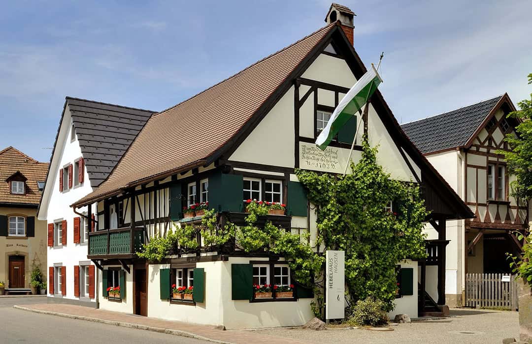 Hausen_im_Wiesental-Hebelhaus_1080 Ein traditionelles Fachwerkhaus mit grünen Fensterläden und Blumenkästen, teilweise mit Efeu bewachsen. Das Gebäude hat ein steiles Dach, eine weiße Flagge und liegt in einer ruhigen Straße in einem europäischen Dorf.