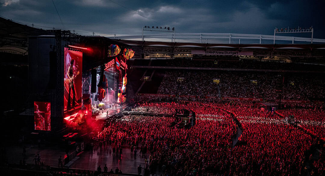 MHP-Konzert_1080 Eine große Menschenmenge füllt in der Abenddämmerung ein Freiluftstadion, das von roten Bühnenlichtern beleuchtet wird, während auf einer hell erleuchteten Bühne eine Band auftritt und auf großen Bildschirmen Nahaufnahmen der Musiker gezeigt werden.