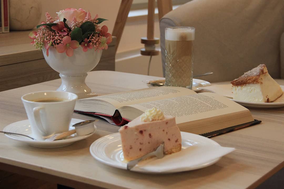 cafetisch_1080 Ein gemütlich gedeckter Tisch mit einem offenen Buch, einer Tasse Kaffee, einem Glas Latte, zwei Stück Kuchen auf Tellern und einem Blumenarrangement in einer weißen Vase. Warme, einladende Raumatmosphäre.