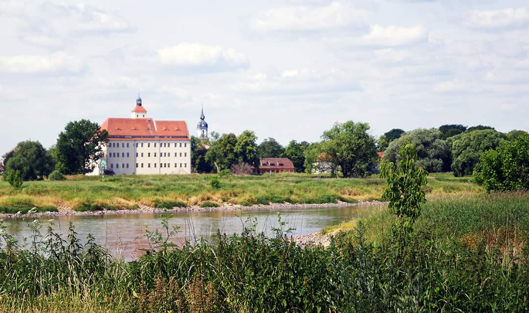 schloss-pertzsch_1080 Ein großes historisches Gebäude mit rotem Dach steht in der Nähe eines Flusses, umgeben von grünem Gras, Bäumen und einem teilweise bewölkten Himmel. In der Ferne hinter den Bäumen ist ein weiterer Turm zu sehen.